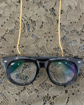 Black acetate eyeglass frames hanging from gold flat lay snake chain glasses accessory over a lace fabric background.