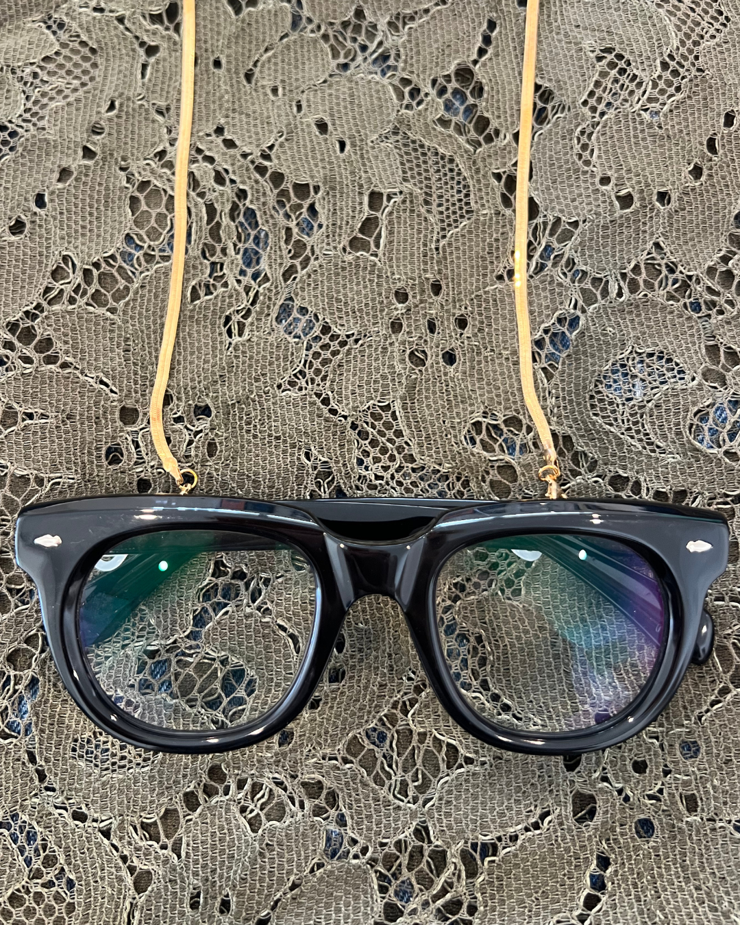 Black acetate eyeglass frames hanging from gold flat lay snake chain glasses accessory over a lace fabric background.