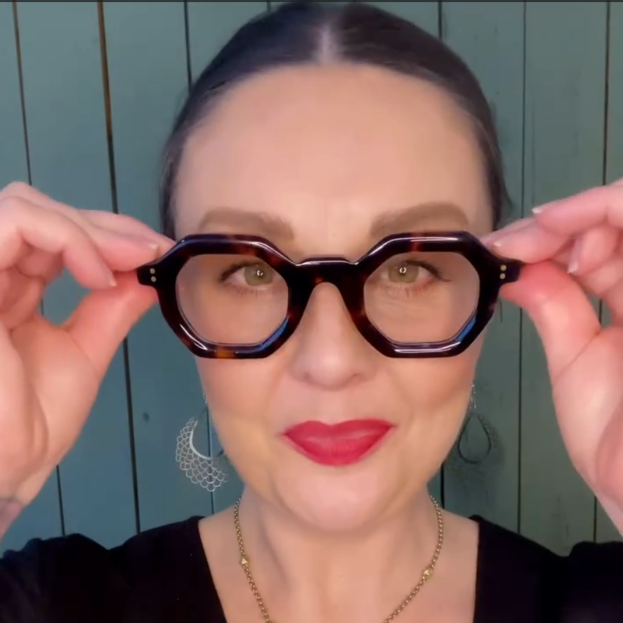 Stylish Australian woman holding up a pair of flattering tortoiseshell reading glasses against a green wooden background - ministry of sight readers