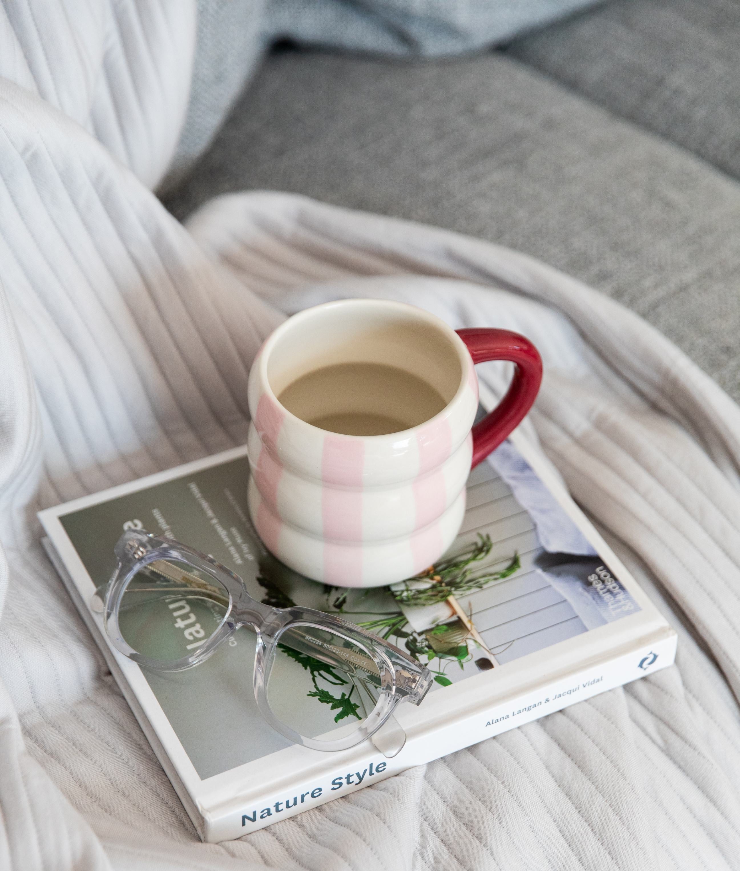 Mug with red handle on a book with clear round stylish reading glasses, surrounded by soft fabric on bed