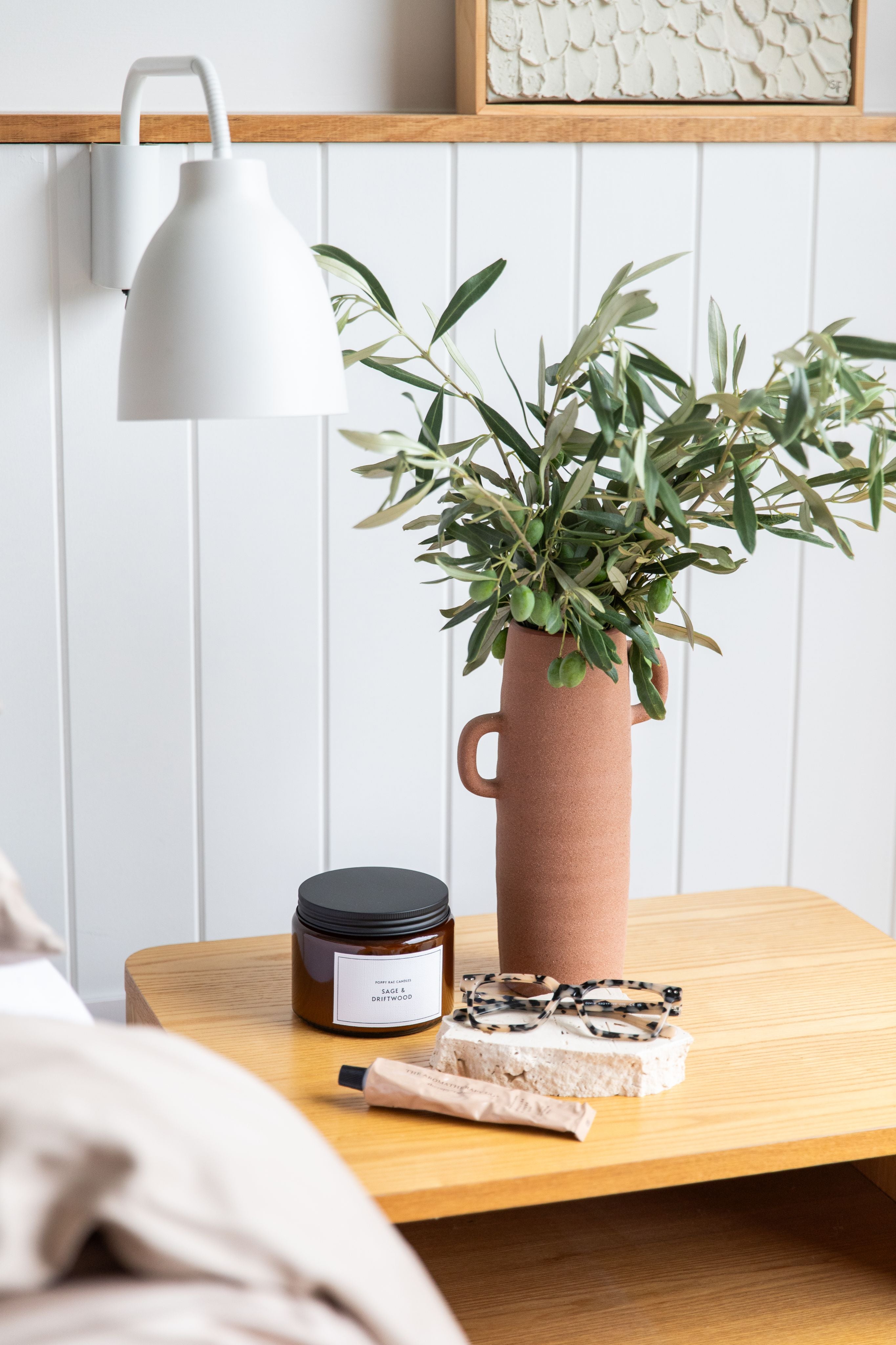 Vase with greenery on a wooden table next to a candle and soap bar, against a white paneled wall. Tortoiseshell reading glasses