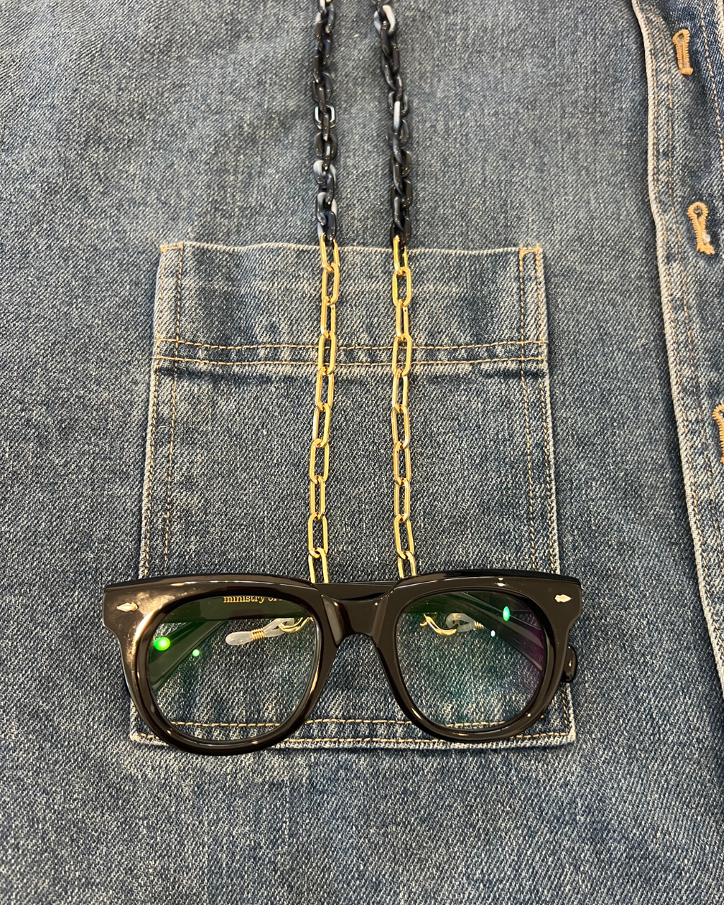 Close-up of a denim jacket with a front pocket, showing gold and navy blue glasses chain to keep your readers close. Look like jewellery. Ministry of Sight. Black oversized round reading glasses. 