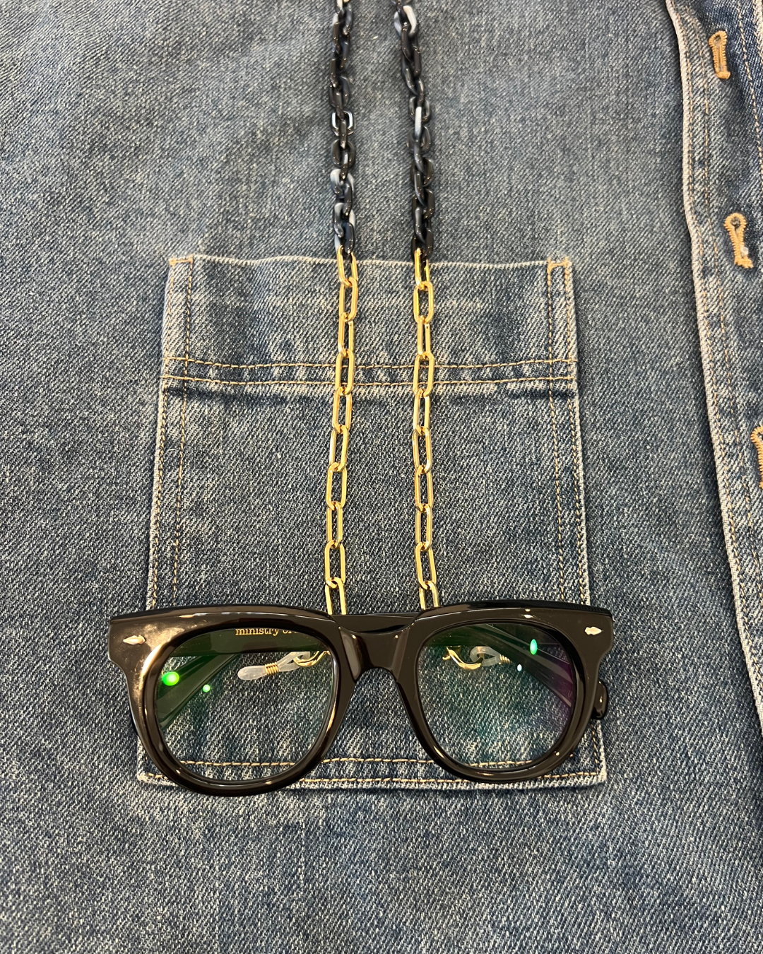 Close-up of a denim jacket with a front pocket, showing gold and navy blue glasses chain to keep your readers close. Look like jewellery. Ministry of Sight. Black oversized round reading glasses. 
