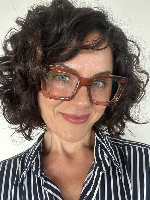 Australian woman over 40 with curly hair wearing stylish pink frame reading glasses and a striped shirt against a plain background - Ministry of Sight