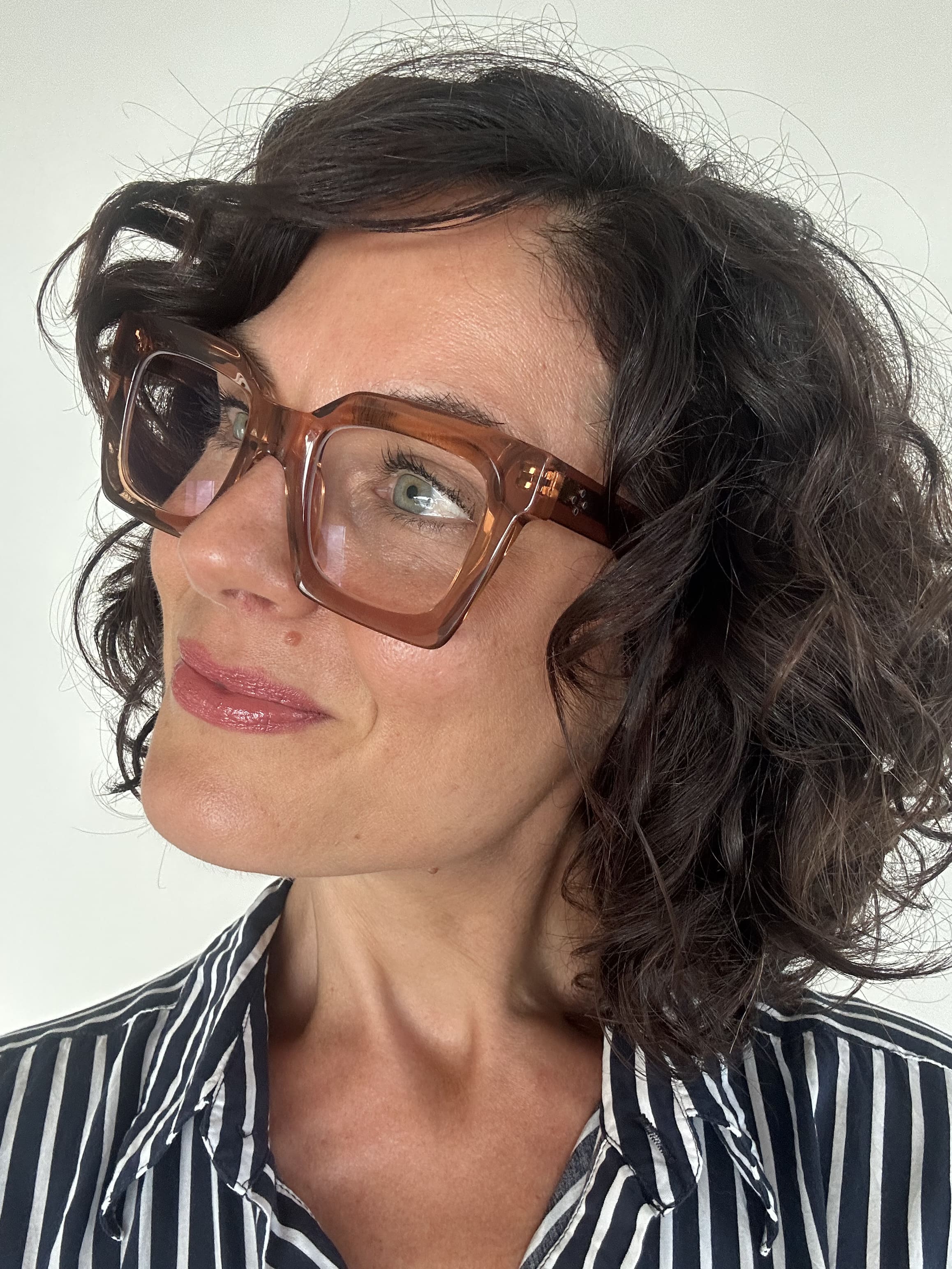 Australian woman over 40 with curly hair wearing stylish clear brown taupe pink frame reading glasses and a striped shirt against a plain background - Ministry of Sight