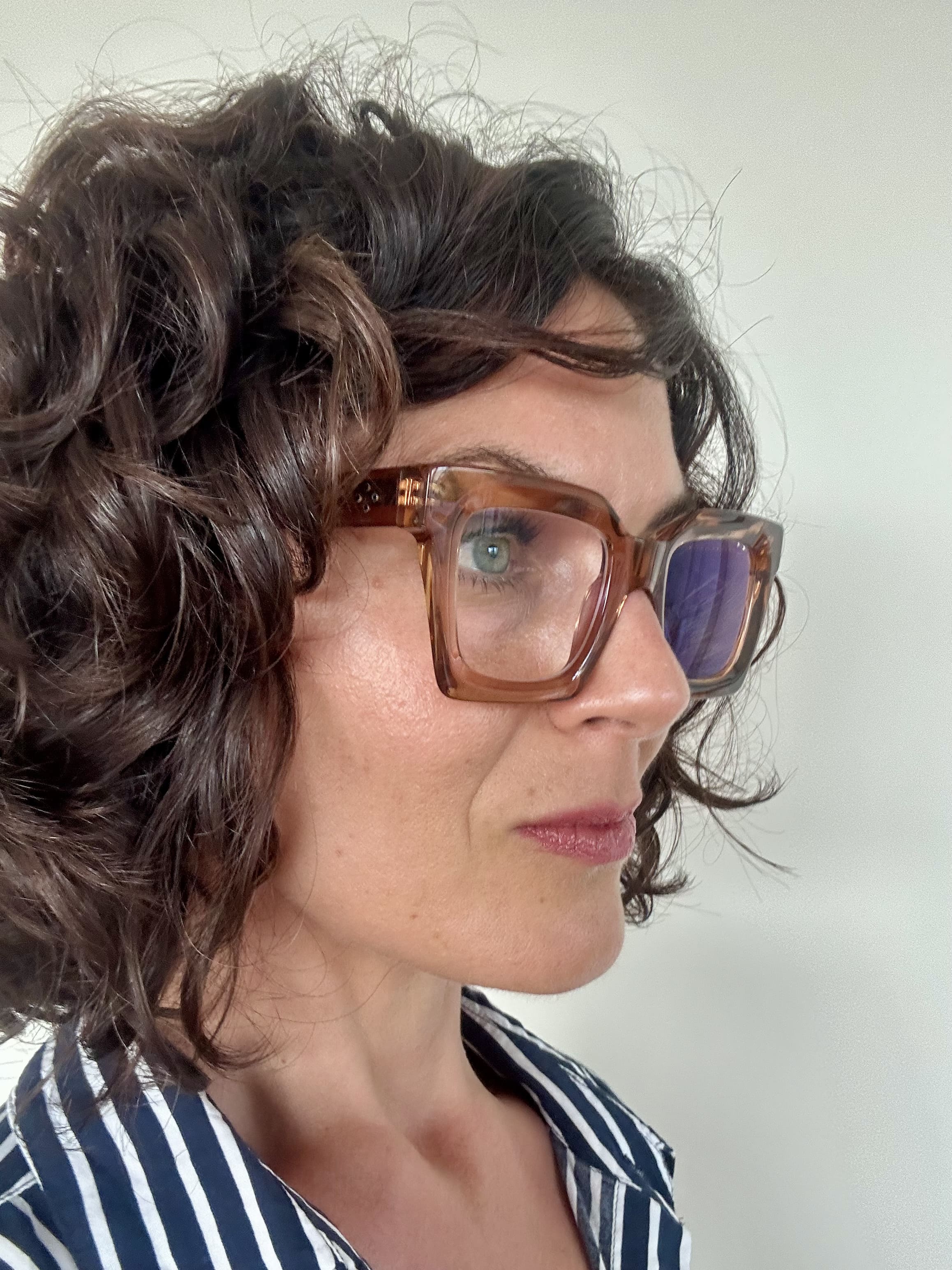 Australian woman over 40 with curly hair wearing stylish pink brown frame reading glasses and a striped shirt against a plain background - Ministry of Sight