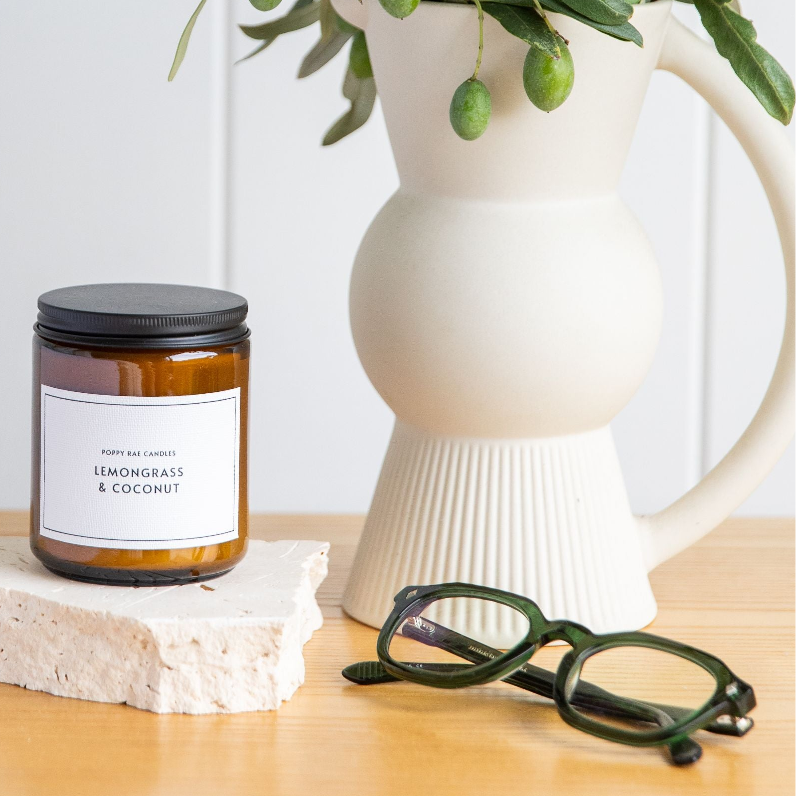 An amber glass jar candle with a black lid and a white label reading 'LEMONGRASS & COCONUT' sits on a light wood table next to a white ceramic pitcher filled with green leaves and a pair of dark green reading glasses in Australia.