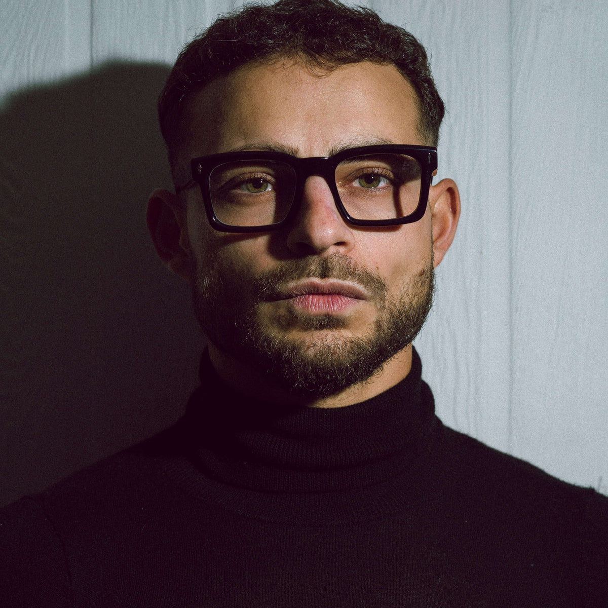 A close-up portrait of a man with short dark hair, a trimmed beard, and thick black rectangular eyeglasses, wearing a black turtleneck sweater against a pale paneled wall.