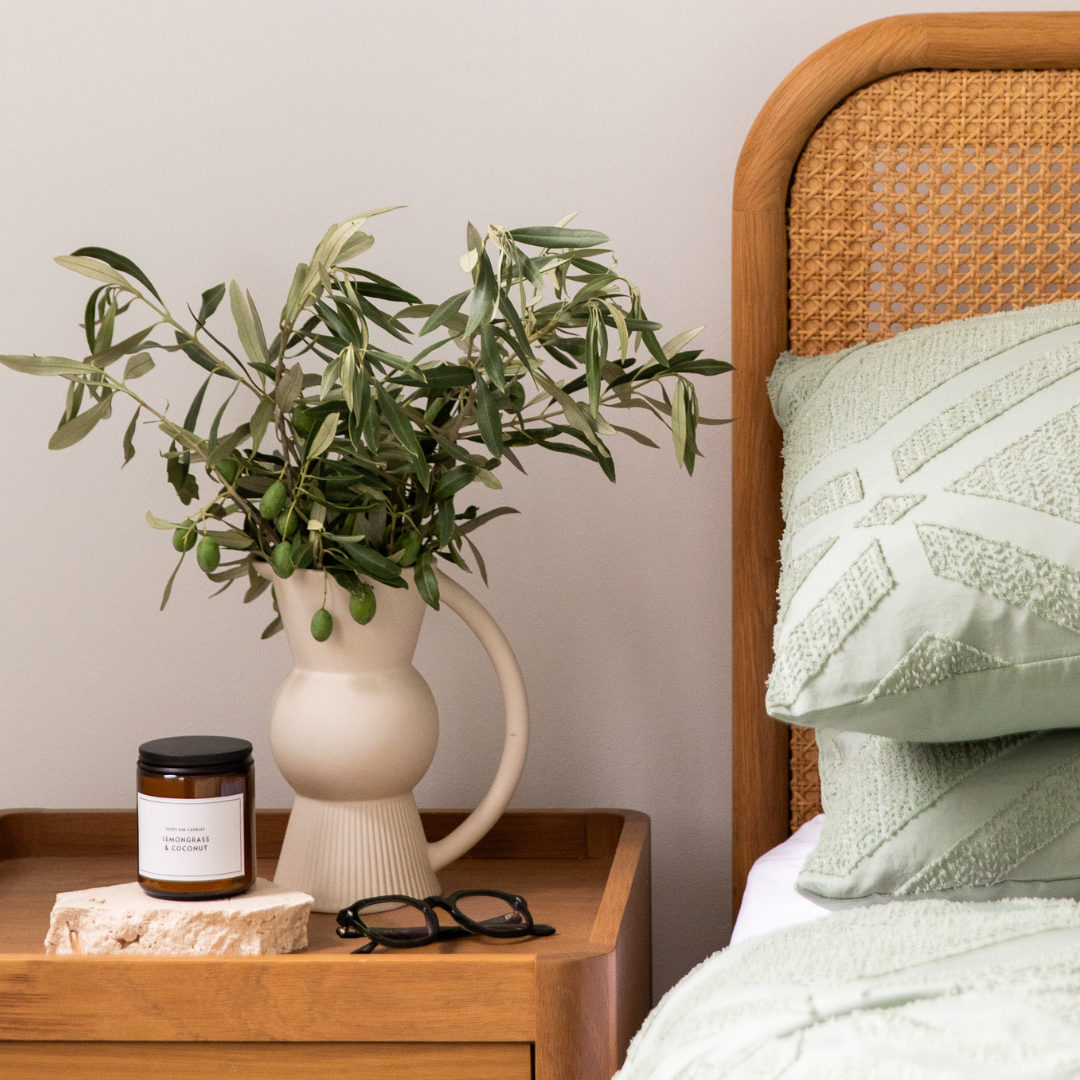 Vase with greenery on a wooden nightstand next to a bed with a green blanket, stylish reading glasses and hand cream.