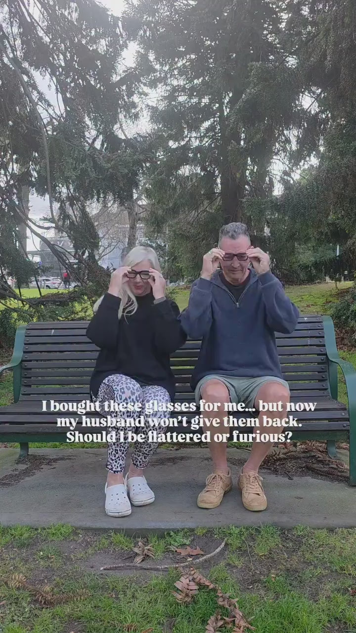 Husband and wife sharing reading glasses on a park bench